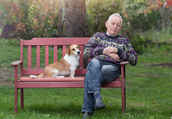 An elderly man sits on a red park bench with his small white and tan dog.