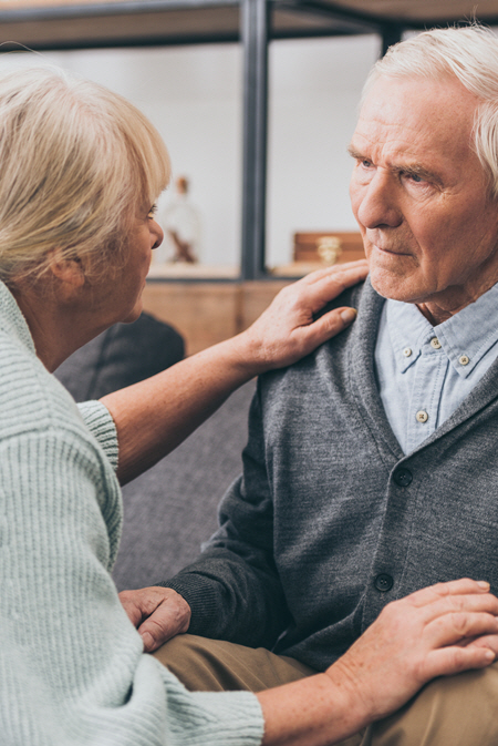 Older woman helping her husband with dementia understand where he is. Her hand is on his shoulder and he looks concerned.