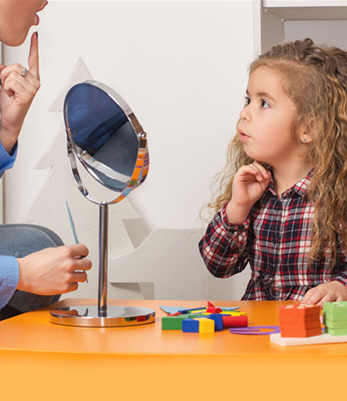 A young girl wearing a plaid shirt works with a speech therapist to pronounce words