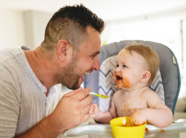 Dad feeding a happy toddler in a high chair out of a yellow bowl