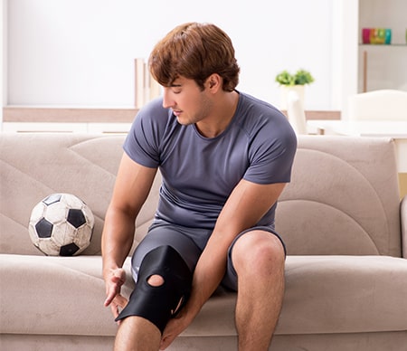 A young man sits on his couch with a knee brace on, his soccer ball sits next to him. He is sad.