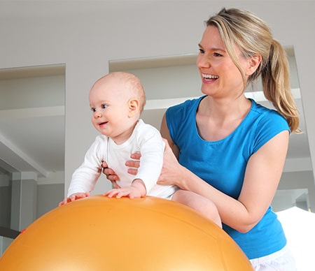 A young mom holds her toddler on a blow up exercise ball. They are both smiling.