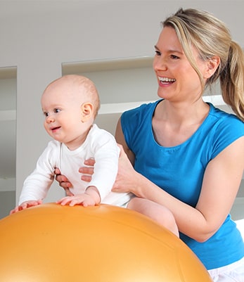 A young mom holds her toddler on a blow up exercise ball. They are both smiling.