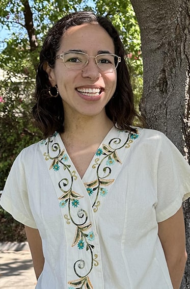 Talia has dark hair and glasses, wearing a cream colored shirt.